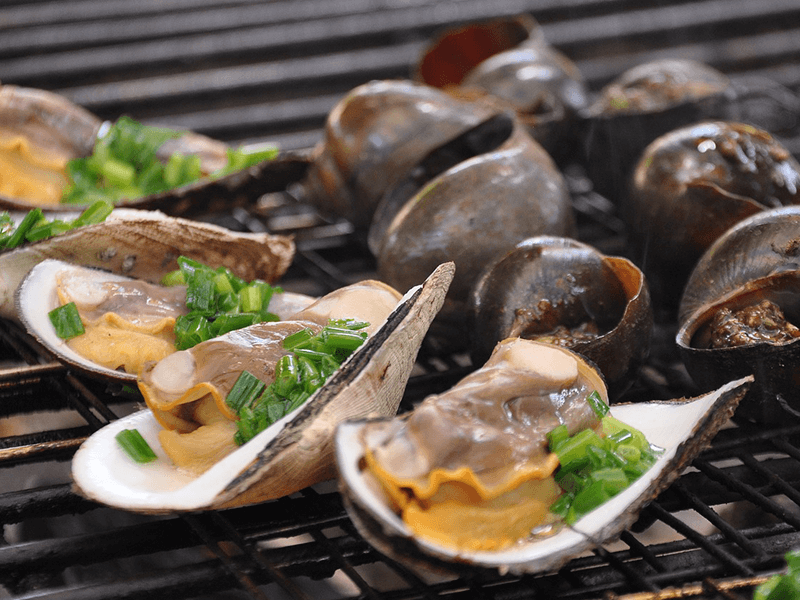 Gegrillte Muscheln und Schnecken auf einem Grillrost, garniert mit gehacktem grünem Zwiebelgemüse. Die Muscheln im Vordergrund zeigen eine saftige Textur, während die Schnecken im Hintergrund dunkel und glänzend sind.
